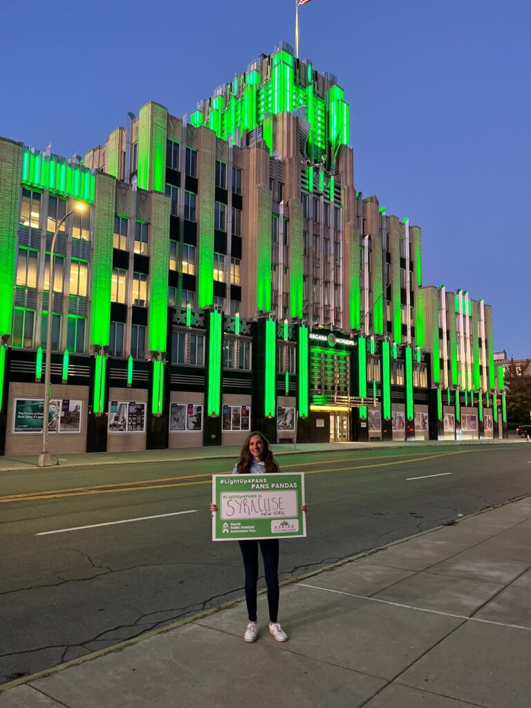 A supporter holds an awareness sign outside the National Grid Building in Syracuse, lit green for Awareness Day 2025