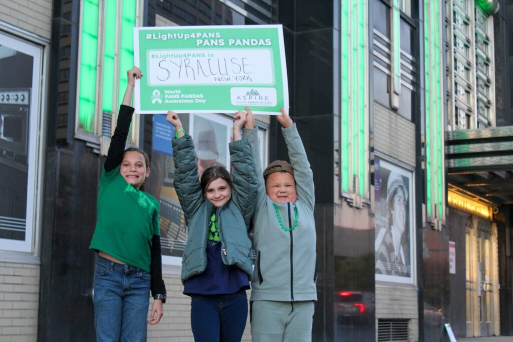 Three children hold a #LightUp4PANS Syracuse sign outside the National Grid Building, lit green for Awareness Day 2025